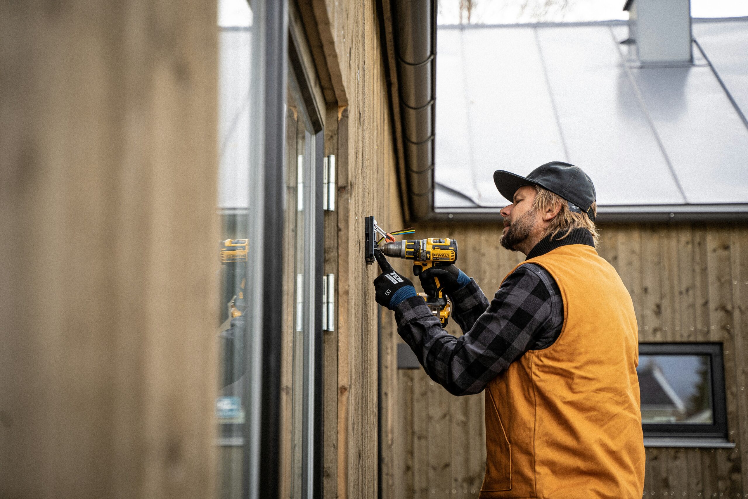 A person wearing a yellow vest and a black cap uses a yellow power drill to install a fixture on the exterior wooden wall of a modern building