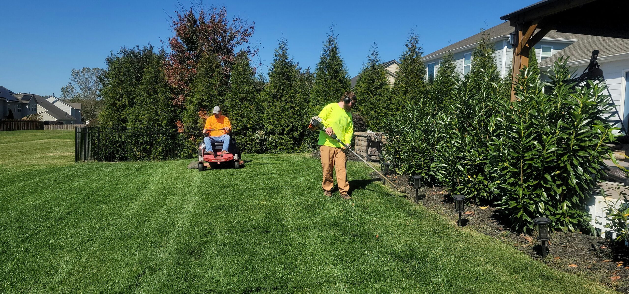 A landscaping crew maintaining a residential backyard on a sunny day. One worker in a neon yellow shirt uses a string trimmer along a garden bed, while another worker in an orange shirt operates a red zero-turn lawn mower in the background