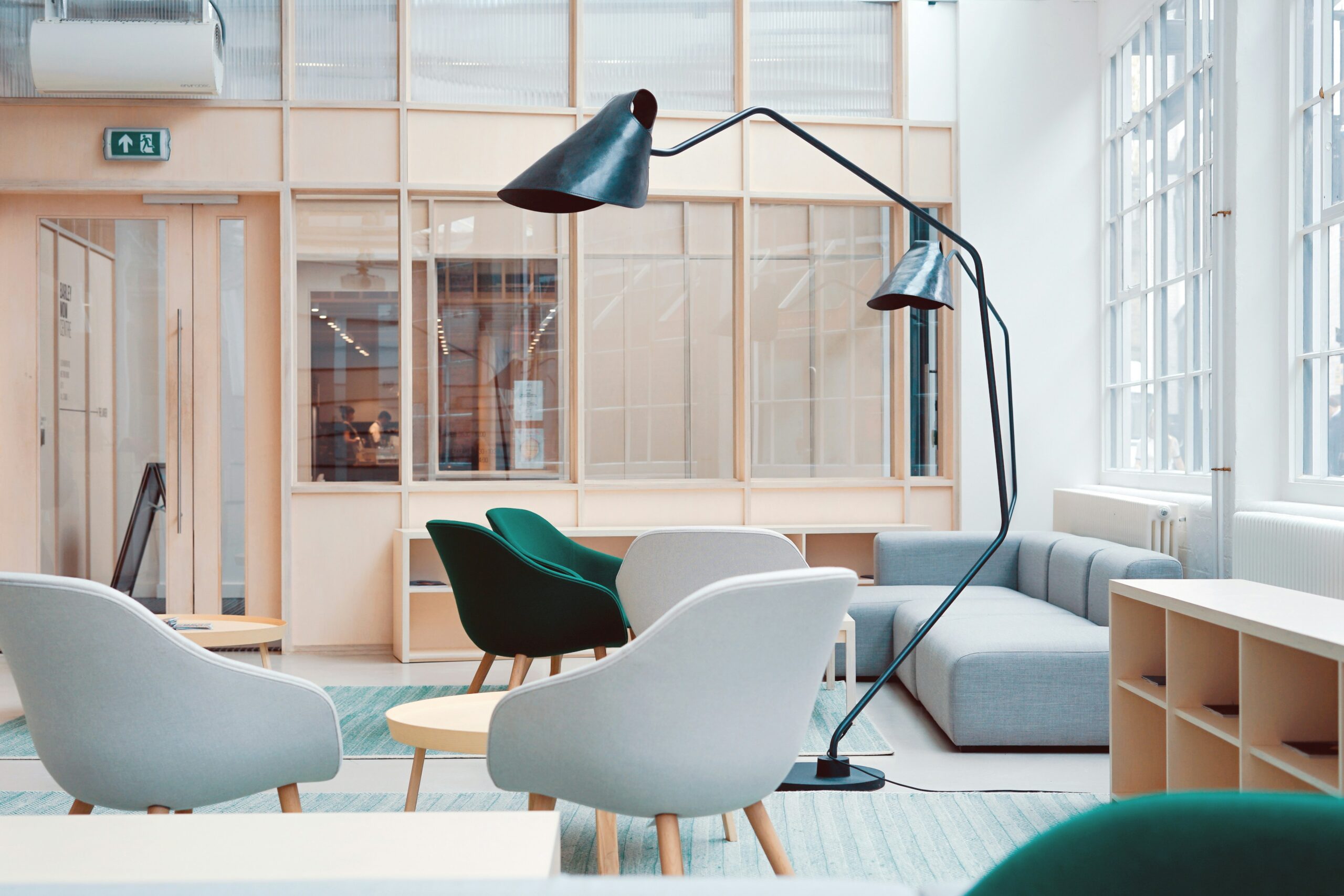 A bright, minimalist modern lounge featuring light gray and forest green armchairs, a gray modular sofa, and a large black multi-arm floor lamp against light wood and glass partitions