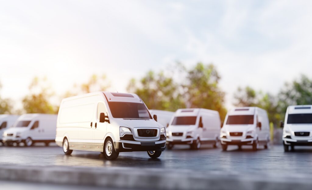 A fleet of white commercial bans parked in a lot with trees in the background.